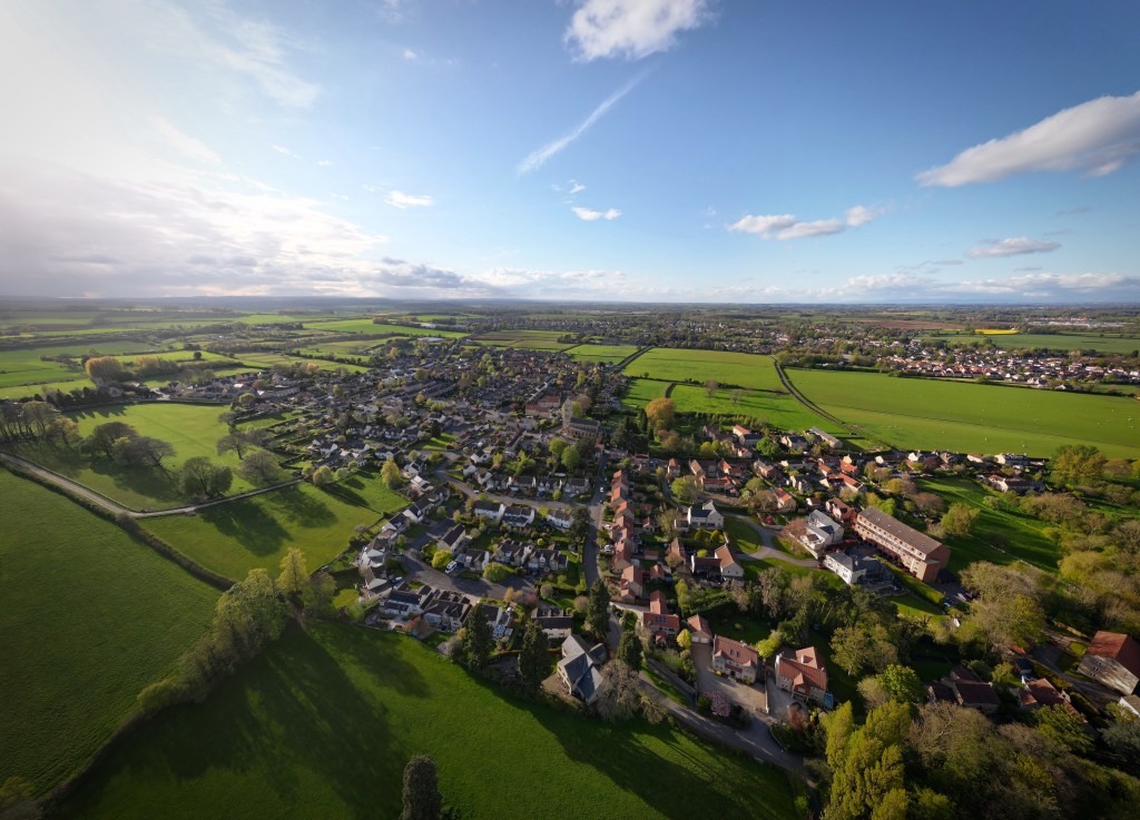 Aerial drone image showing a scenic village landscape in West Yorkshire, with rooftops, greenery, and countryside views captured from above.